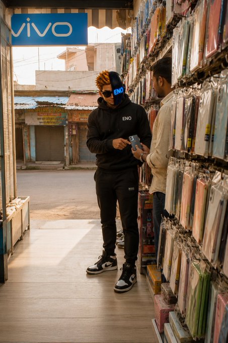 A young man wearing a black 'ENO' hoodie and a blue and black mask stands in a mobile phone accessories shop looking at his phone, with a 'vivo' sign visible in the background.