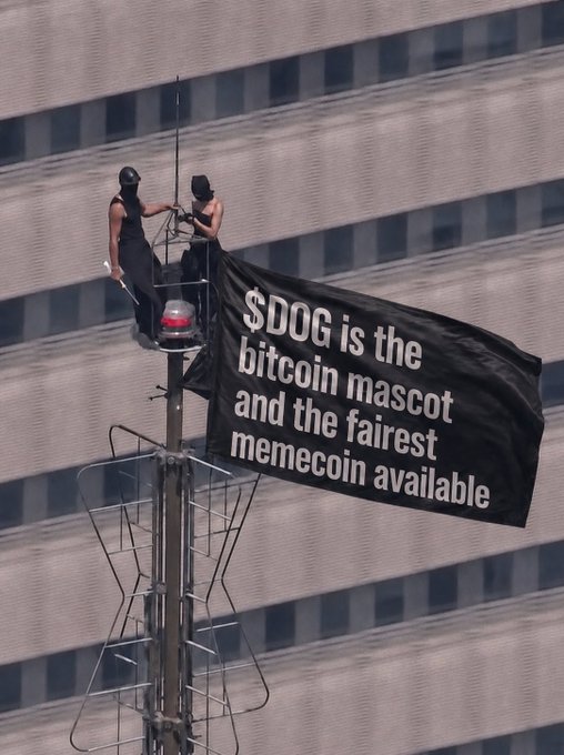 Two masked individuals hang a black banner from atop a structure against a building facade, reading '$DOG is the bitcoin mascot and the fairest memecoin available.'