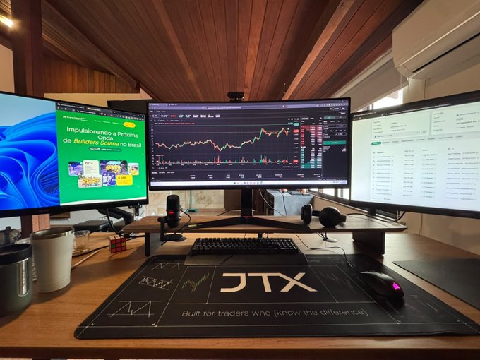 A multi-monitor trading setup on a wooden desk showing crypto charts and portfolios, featuring a JTX desk mat, mechanical keyboard, mouse, and a microphone.
