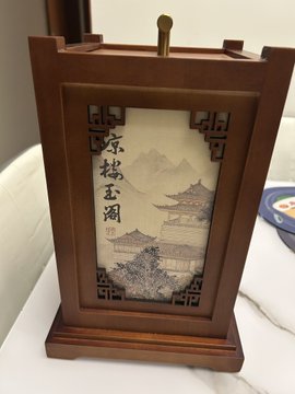 The image features a wooden box with an oriental painting depicting a temple nestled among mountains. The box has a gold-colored lid that adds elegance to its rustic appearance. The painting showcases intricate details of the temple, including trees and mountains in shades of green and brown. The box is placed on a white surface, which contrasts beautifully with the wooden finish and enhances the colors of the painting.