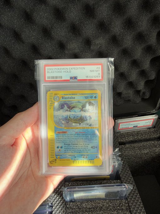 A hand holds a PSA 8 graded 2003 Pokémon Expedition #4 Blastoise Holo card (cert #151323285) in front of a foam-lined case containing other graded cards.