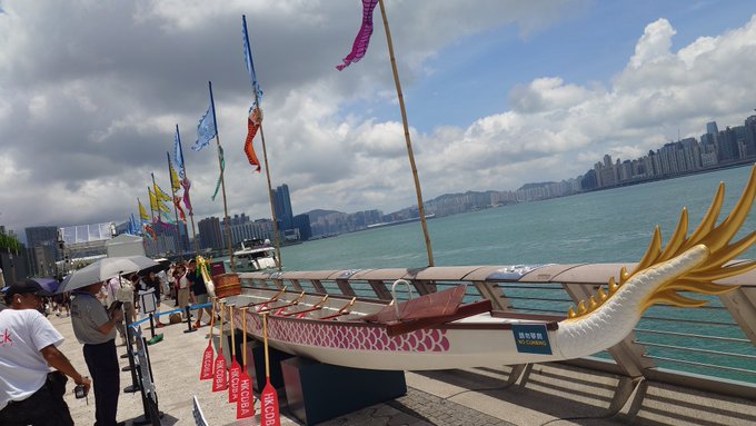 The image features a large dragon boat with a gold fin, located near the water's edge. The boat has red and white stripes along its sides, giving it an eye-catching appearance. There are several people standing around the boat, possibly admiring or preparing for a rowing event. In addition to the dragon boat, there is also a small boat visible in the background on the right side of the image.

The post accompanying the image provides information about the dragon boat and its location. It reads: "6/20 中央香港  dragonsong 中央香港  dragonsong #stepn #stepngo". This suggests that the dragon boat is located in a city near the sea, possibly in Hong Kong or another coastal city.