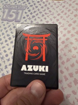 A hand holding a black 'Azuki Trading Card Game' card with a red Torii gate design, shown against a patterned background.