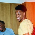 A casual photo of a smiling young man in a white t-shirt standing next to another person in a room with wooden cabinets.