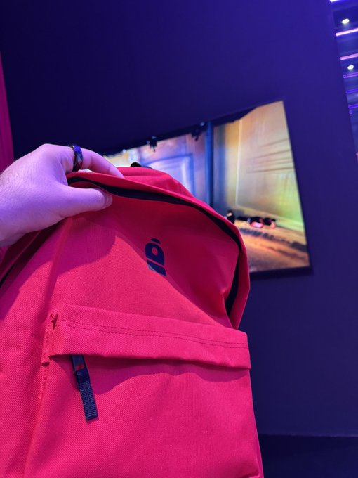 Close-up of a hand holding a red backpack with '100' logo in a dark room with purple/blue lighting and a screen displaying what appears to be a racing game.