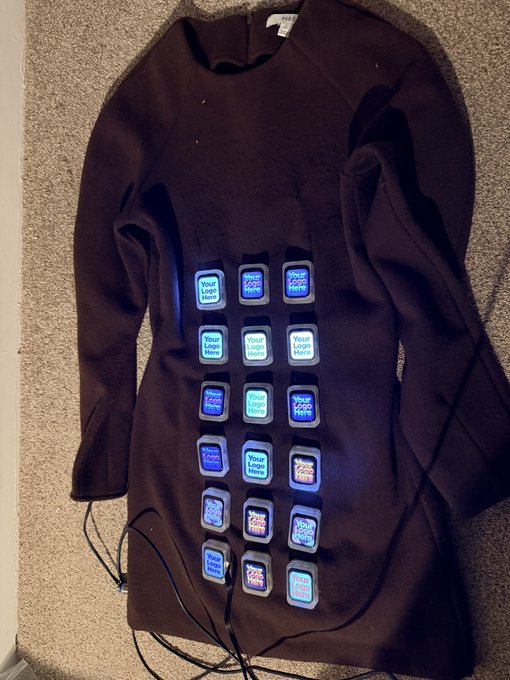 A black long-sleeve dress lies on a carpeted floor with multiple small screens attached to the front, each displaying scrolling placeholder text like 'Your Logo Here' in blue.