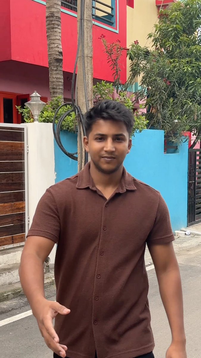 A young man in a brown short-sleeve button-up shirt stands on a street in front of a colorful pink and blue building with trees in the background.