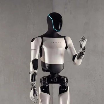 A humanoid robot with a Tesla logo on its chest, featuring a white and black design with a glowing cyan visor, standing against a gray background.