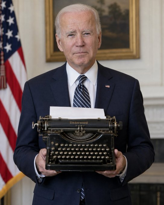 President Joe Biden in a dark suit stands in front of an American flag and fireplace, holding a vintage Underwood typewriter with paper loaded.
