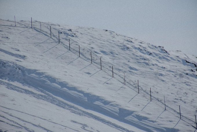 A snow-covered hillside with a wire fence supported by wooden posts running diagonally across the slope.