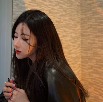 A close-up profile portrait of a young woman with long dark hair and red lipstick, looking downward against a textured wall background.