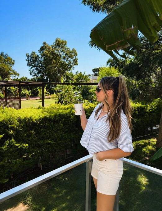 A young woman with long blonde hair wearing sunglasses, a blue and white striped shirt, and white shorts stands on a balcony holding a drink, with a tropical garden in the background.