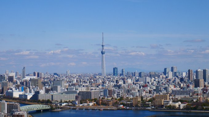 A wide daytime cityscape of Tokyo featuring the Tokyo Skytree tower prominently in the center, with the Sumida River visible in the foreground.