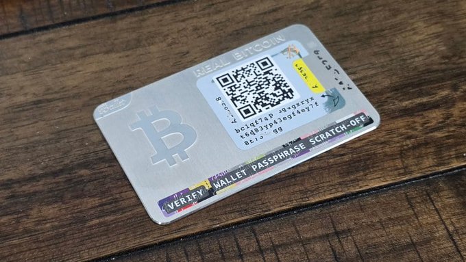 A metal Bitcoin cold wallet card with a QR code and a scratch-off panel for a wallet passphrase, resting on a wooden surface.