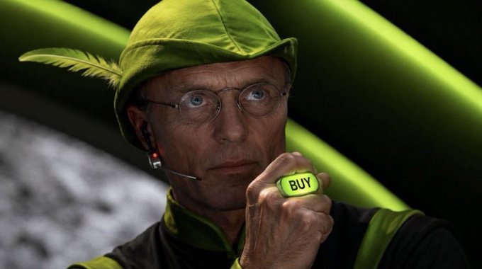 A man wearing a green jester-style hat with a feather and round glasses, holding a glowing green 'BUY' button near his face against a green-toned background.