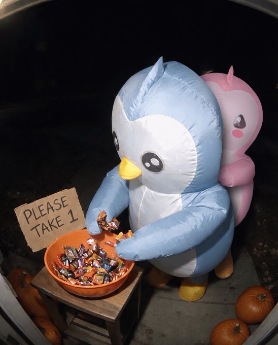 An inflatable penguin mascot distributes Halloween candy from an orange bowl, accompanied by a cardboard sign reading "PLEASE TAKE 1." Pumpkins scattered at its feet and a pink plush companion set the festive scene, teasing tomorrow's Halloween costume reveal.