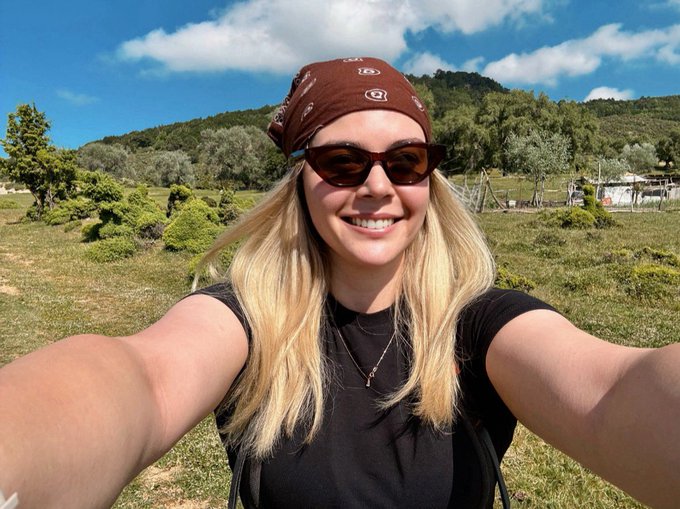 A smiling woman with long blonde hair, wearing a brown bandana and sunglasses, takes a selfie outdoors against a backdrop of green hills and a partly cloudy blue sky. The casual, adventurous outdoor setting contrasts with the post's focus on data-driven forecasting from ARC Intelligence, possibly suggesting she's taking a break from analyzing the platform's uncertainty bands.