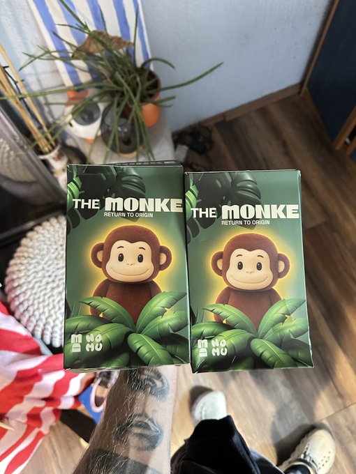 Two green boxes of 'THE MONKE - Return to Origin' collectible figures featuring a cartoon monkey character with a glowing halo, displayed on a table with plants in the background.