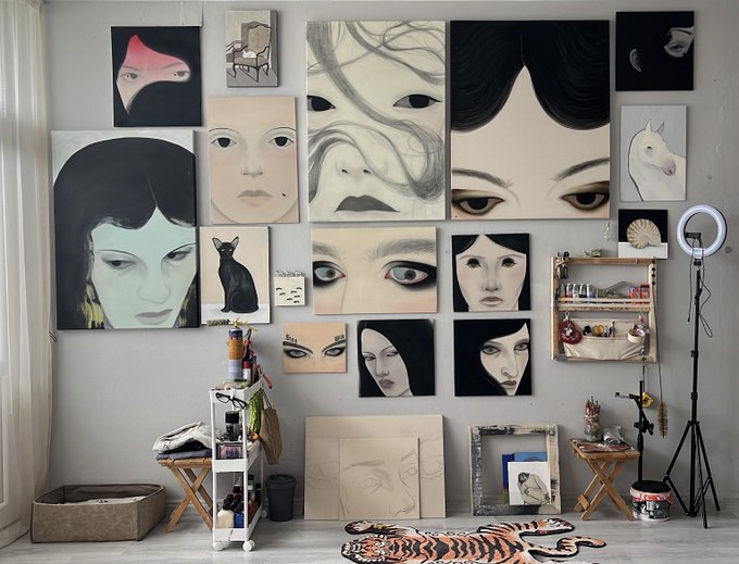 An artist's studio wall displays a curated gallery of portrait paintings featuring faces and a black cat, accompanied by art supplies and a ring light on a tripod.