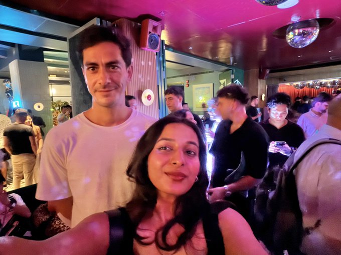 The image shows a young woman taking a selfie at a vibrant, crowded event with pink and purple lighting, posing alongside a tall young man in a white t-shirt—likely at the TOKEN2049 "bingbong party" mentioned in the caption, where she briefly met and was starstruck by him in person.