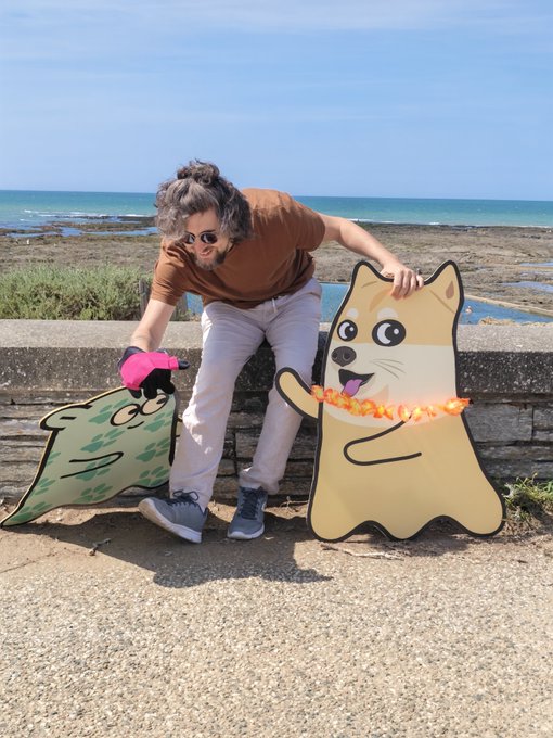 A person wearing a brown shirt, sunglasses, and a pink glove poses outdoors by the sea alongside two cartoon-style wooden cutouts of a green creature and a Shiba Inu dog.
