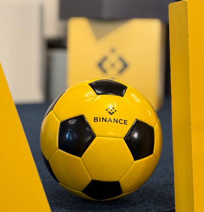 The image features a yellow soccer ball with black spots, which appears to be a regulation size soccer ball. It is placed on a blue carpeted floor, surrounded by various objects such as a box, a chair, and a book. The background of the image includes a yellow wall and a gray wall, creating an interesting contrast between the colors in the foreground and background. The text "Every season is Binance season" can be seen in the image, which seems to be related to the soccer ball or the post it appears to be connected to.