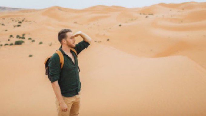 A man wearing a green shirt and backpack stands in a vast desert, shielding his eyes as he scans the arid landscape. The image visually echoes the "Oasis or die" sentiment, capturing a traveler searching desperately for water in an endless sea of sand.