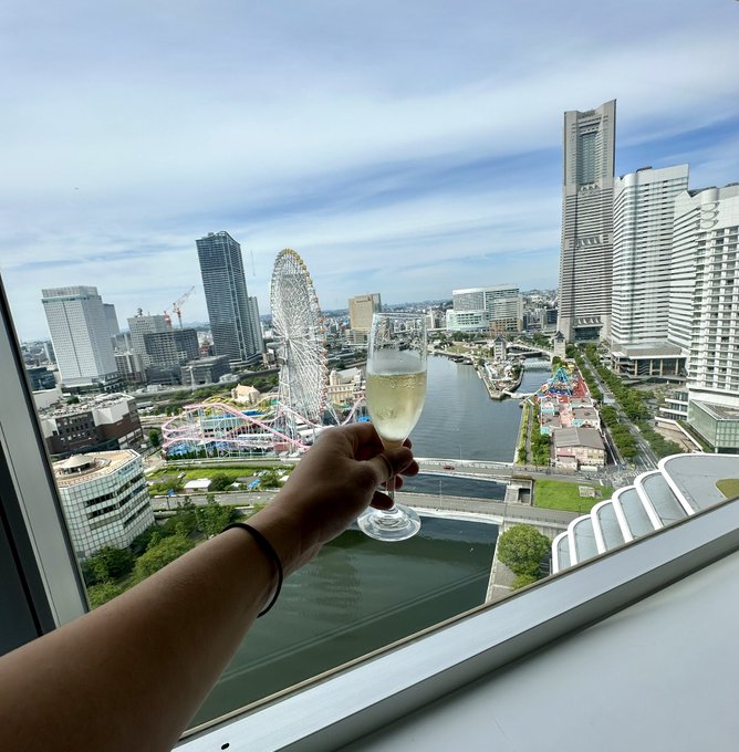 A hand holding a champagne flute toasts toward a window view of a city skyline featuring a Ferris wheel, waterfront, and tall skyscrapers including one with 'ANA' signage.