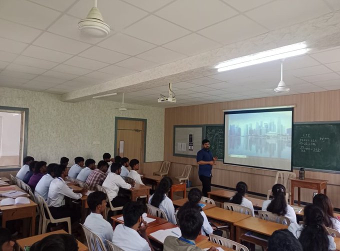 A classroom scene showing a teacher standing in front of a projected cityscape image while students seated at wooden desks watch the presentation.
