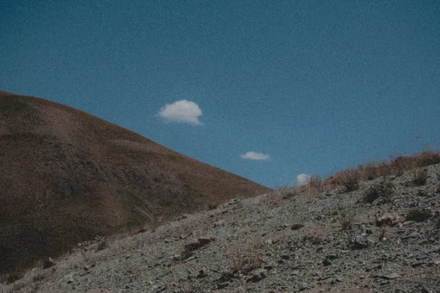 A grainy photograph of a barren, rocky hillside under a deep blue sky with a single small white cloud, featuring visible text '3A' in the corner.