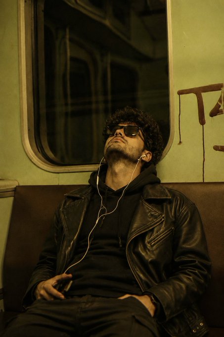 A young man with curly hair and sunglasses dozes off on a dimly lit train, his head tilted back as earbuds play music and his hand loosely grips a phone. The weary, introspective pose echoes the post's late-night grind mindset, capturing a quiet moment of exhaustion amid the hustle of building and shipping.