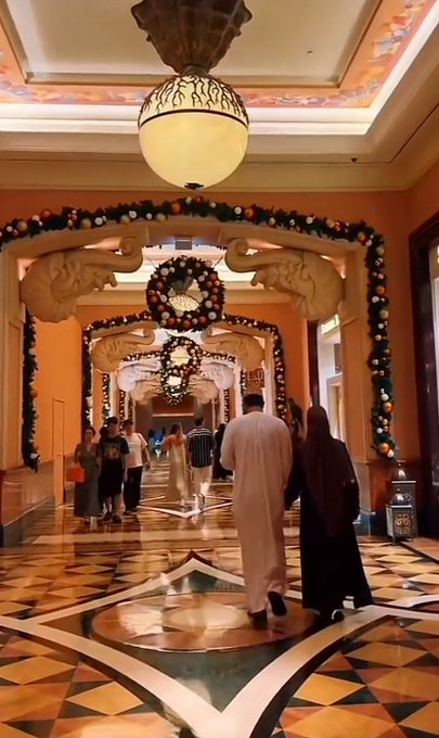A luxurious hotel hallway adorned with elaborate floral arches, ornate elephant sculptures, and decorative pendant lighting, where guests stroll along a patterned marble floor—likely the Atlantis resort in Dubai, a popular destination for crypto influencers and lifestyle content like this GM (Good Morning) post.