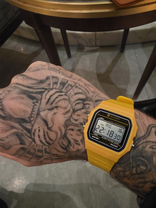 The image features a person wearing a yellow wristwatch with black numbers and hands. The watch has a tattoo of a tiger's face on their wrist. The background shows a dining table with chairs, suggesting that this scene takes place at a restaurant or similar setting. A caption in the image reads "Turns out Mert himself approved the Blockspäti @Zcash watches." This indicates that the person wearing the watch has received approval from someone named Mert for their choice of wristwatch.