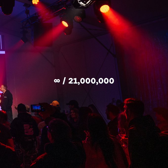 A photo taken inside a venue with red stage lighting, showing a crowd of people and an infinity symbol followed by '/ 21,000,000' superimposed in white text.