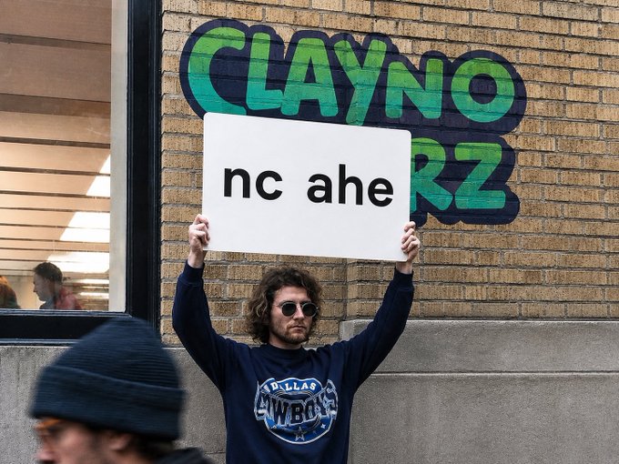 A man with curly hair and sunglasses holds up a white sign reading 'nc ahe' in front of a brick wall with graffiti-style lettering spelling 'CLAYNO' and 'RZ'.