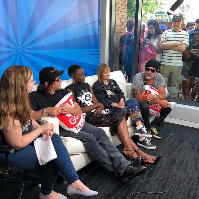 A group of actors, including Norman Reedus wearing a "WIDOW" branded cap, sit on a white couch during a panel interview, with a crowd of fans visible through the window behind them, capturing a candid moment from San Diego Comic-Con.