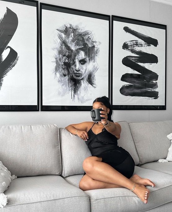 A woman in a black dress sits on a light gray sofa holding a dark mug, with three large black-and-white abstract art pieces hanging on the wall behind her.