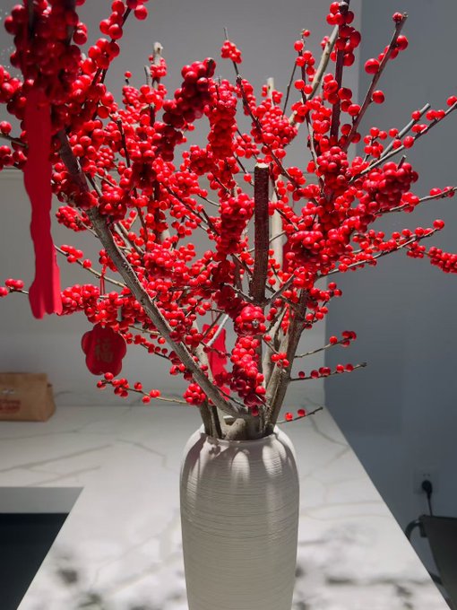 Decorative arrangement of red berries (likely winterberry or holly branches) in a textured silver/pewter vase on a marble countertop, with small red decorative tags hanging from the branches.
