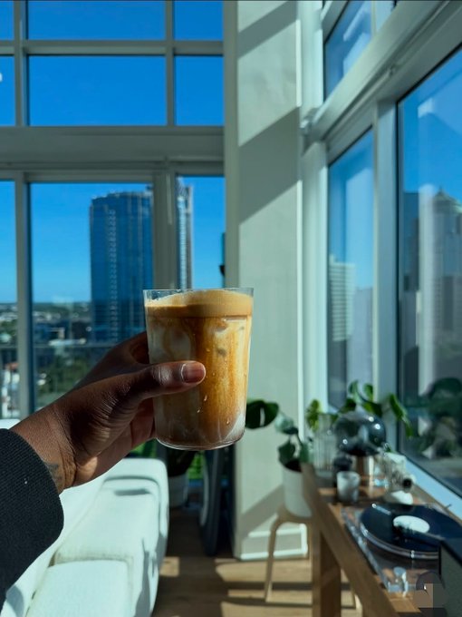 A hand holds up a glass of iced coffee against a bright window with a cityscape view, set in a sunlit apartment with plants and a record player visible.