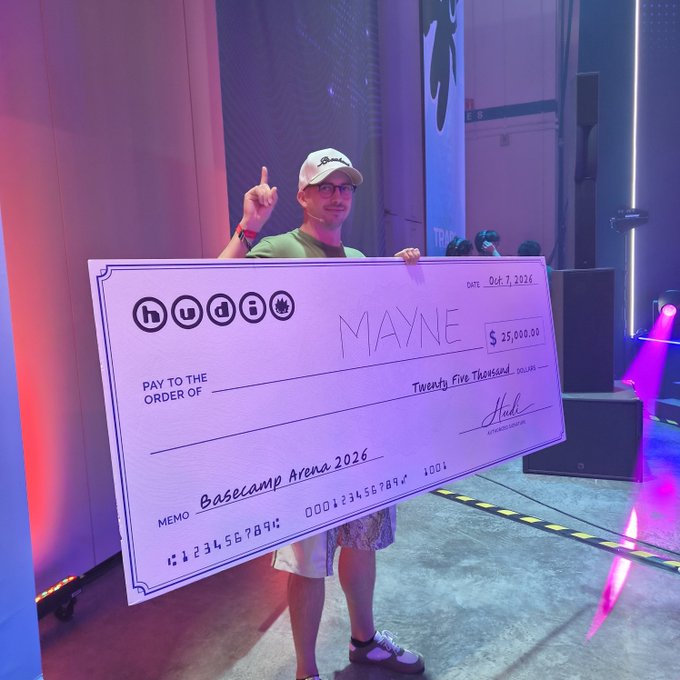 A man wearing a green shirt and white cap holds an oversized novelty check made out to 'MAYNE' for $25,000.00 from Hudi, dated Oct. 7, 2026, with memo 'Basecamp Arena 2026'.