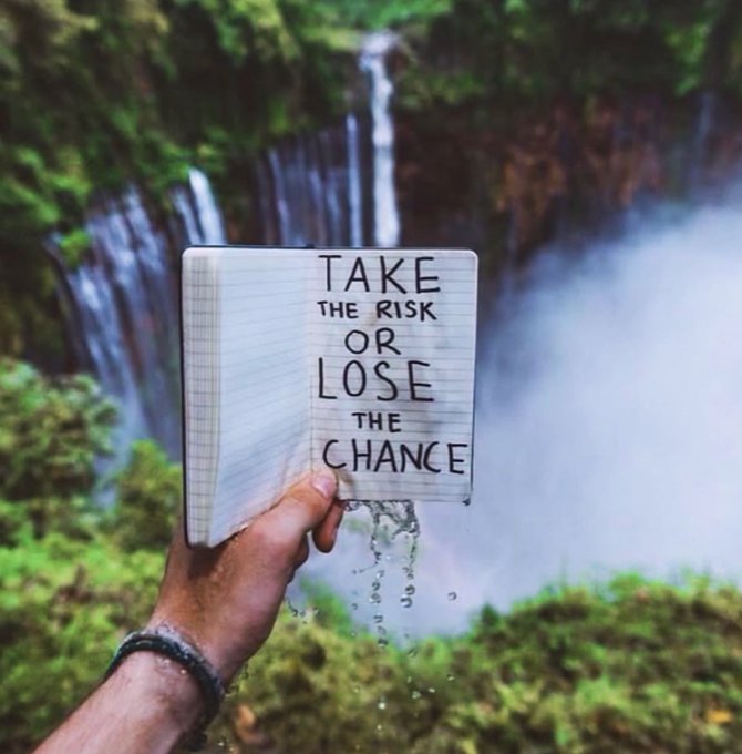 An inspirational photo of a hand holding open a lined notebook in front of a misty waterfall, with the handwritten motivational message 'TAKE THE RISK OR LOSE THE CHANCE' on the page.