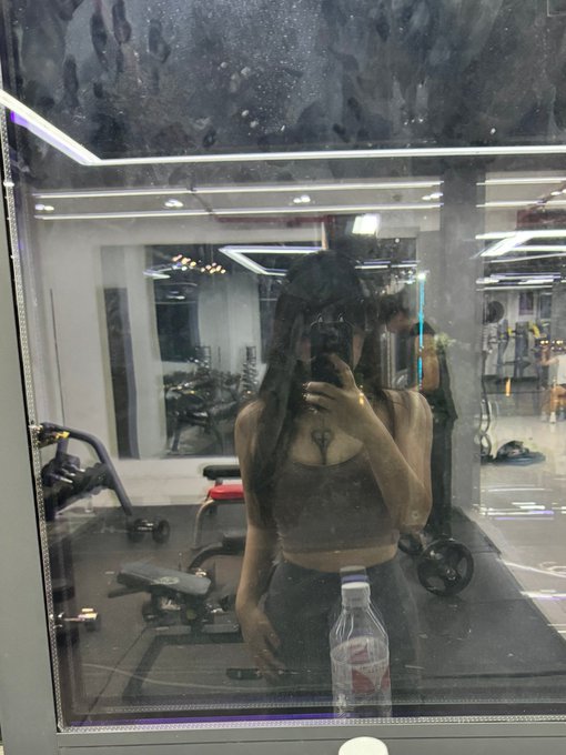 A woman takes a mirror selfie at the gym holding a water bottle and phone, fitting the caption's message about balancing weekend rest with gym workouts to stay focused for finding the next "100x gem" in crypto/trading.