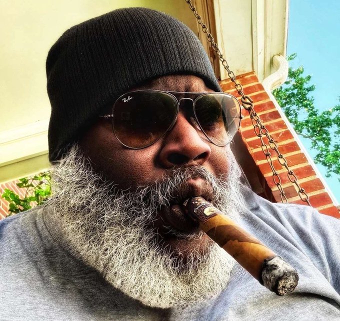 A close-up portrait of a man wearing a black beanie and Ray-Ban aviator sunglasses, with a long gray beard, smoking a cigar while sitting on what appears to be a porch swing.