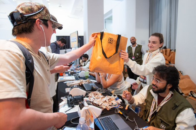 The image shows a group of people gathered around a table, with one person holding an orange bag over another's shoulder. There are also two laptops visible on the table, suggesting that they might be working on a project or discussing ideas related to technology. A water bottle is placed nearby as well. The post about hackers and swag bags seems to be promoting a specific event or gathering for tech enthusiasts in New York City.