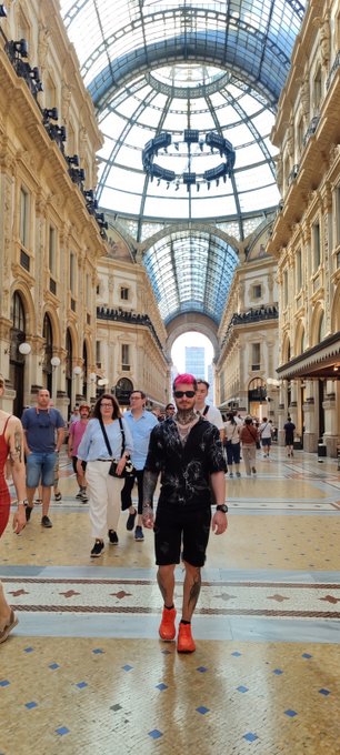 A person with pink hair, sunglasses, a black patterned shirt, black shorts, and red sneakers posing for a photo inside a grand, ornate shopping arcade with a stunning glass dome ceiling.