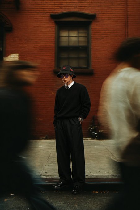 A street style photograph of a person in all-black attire including a bucket hat with red details, standing against a red brick building while blurred figures move past in the foreground.