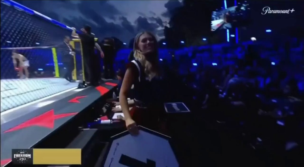 The image shows a woman standing next to a large screen TV with a caption that reads " UFC Freedom 250 from crypto to the UFC octagon". The scene takes place at an event where the UFC has been featured, possibly as part of a promotional campaign or special edition. The woman is holding up a sign and smiling for the camera, likely promoting the event or the UFC's involvement in it.