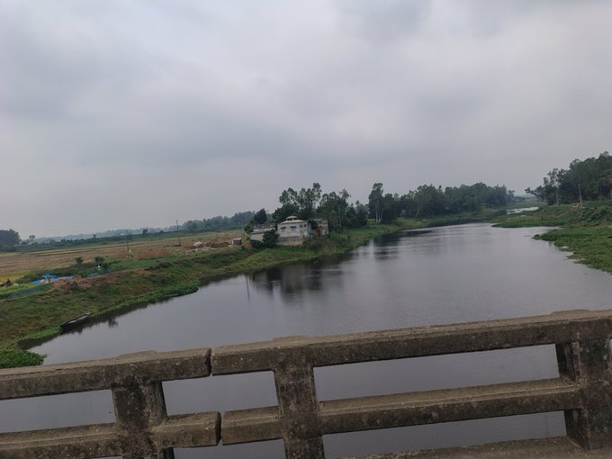 The image shows a river flowing through a rural area, surrounded by trees and fields. The sky above appears cloudy, suggesting an overcast day. In the foreground of the photo, there's a concrete bridge spanning across the river, with a railing on one side. A small house can be seen in the background near the water's edge. The post accompanying this image is about generating activity for Nomisma Network BULLISH and uses the @NomismaNetwork BULLISH tag to describe its content.
