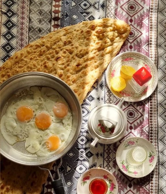 A top-down photo shows a traditional Persian breakfast spread with flatbread, eggs in a pan, tea, and jam served on patterned rugs and floral dishes.