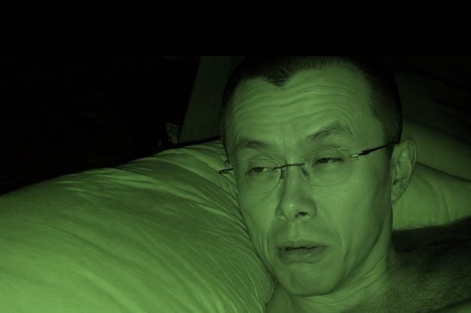 A man with glasses lies on a pillow in darkness, captured with night vision that gives the scene a green tint.